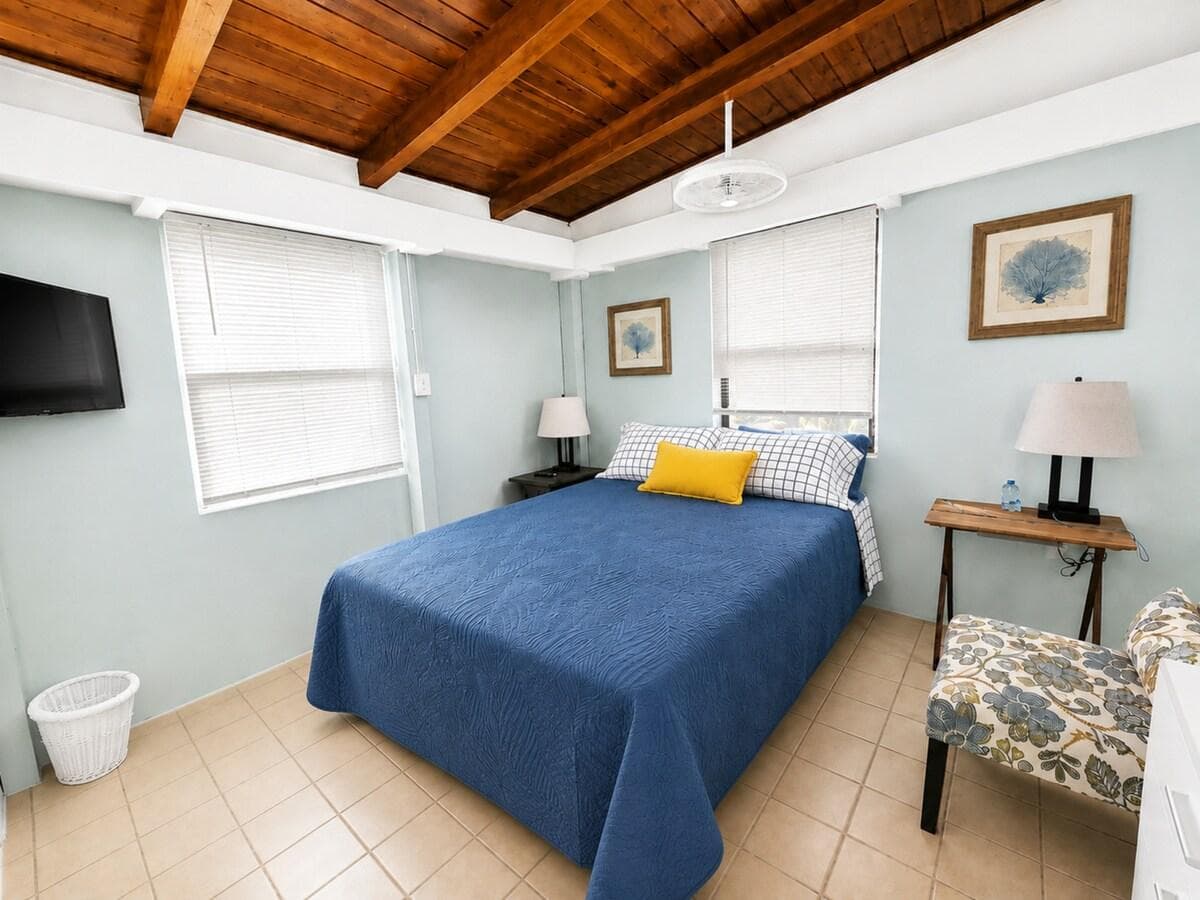 Bright and inviting coastal bedroom featuring a comfortable bed with crisp blue bedding, charming beach-inspired décor, and a beautiful exposed wood-beam ceiling. A relaxing retreat to unwind after a day at the beach.