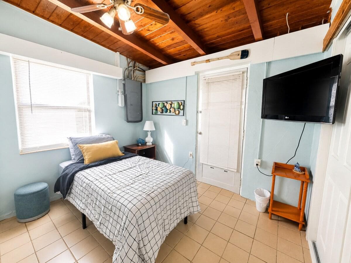 Cozy and inviting bedroom featuring comfortable bedding, a wall-mounted TV, and charming coastal décor. The beautiful exposed wood-beam ceiling adds warmth and character, while plenty of natural light creates a bright, relaxing space to unwind after