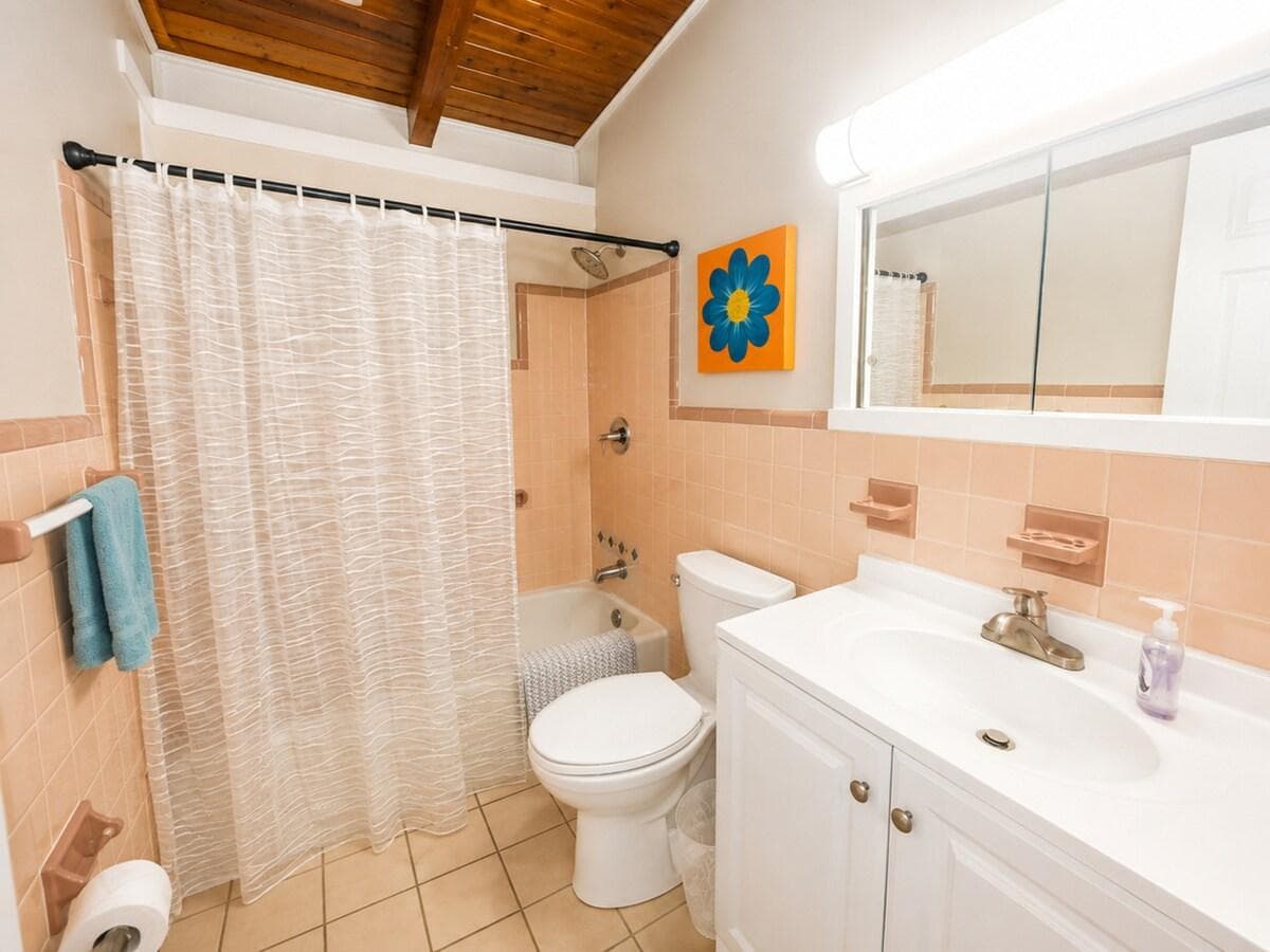 Bright, clean bathroom featuring a full tub and shower, spacious vanity, and charming coastal touches. Warm wood ceiling details and cheerful décor create a comfortable, inviting space to refresh after a day at the beach.